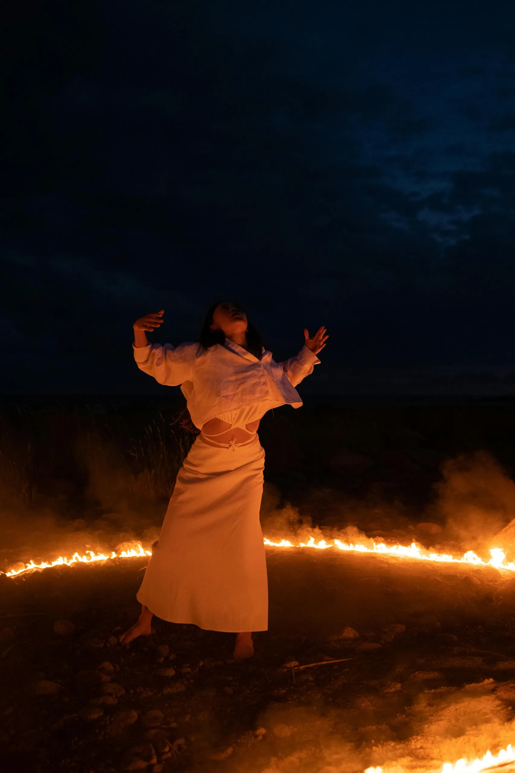 A woman performs an enchanting ritual within a ring of fire under a dark night sky, evoking mystical allure.