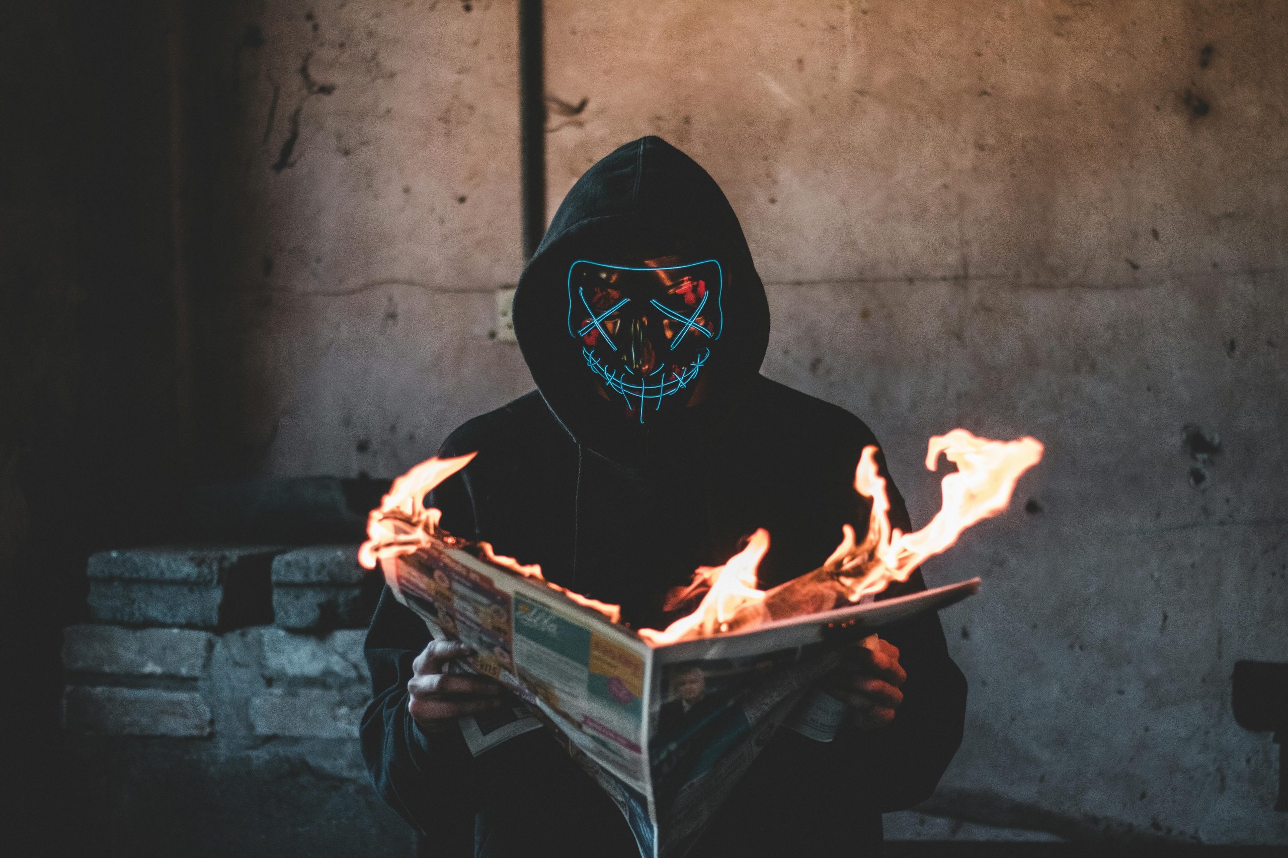 A hooded figure with a glowing mask holds a burning newspaper inside a dim room.