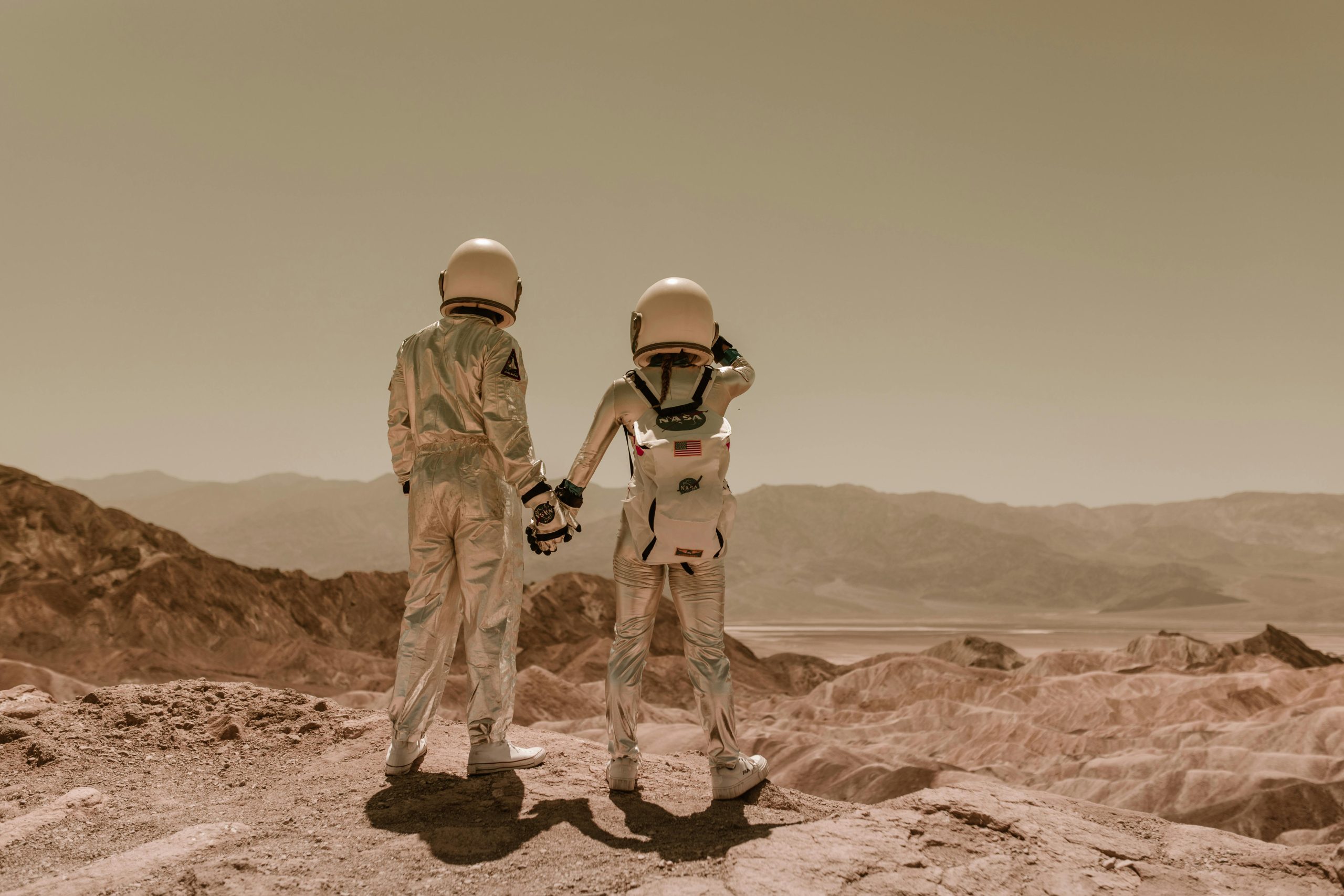 Two astronauts in space suits holding hands in a desert, Mars-like terrain.